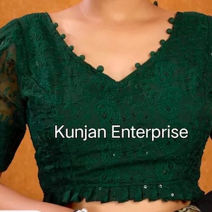 May include: A green embroidered blouse with a V-neckline and short sleeves. The blouse has a ruffled hem and is decorated with small, white buttons. The text "Kunjan Enterprise" is visible on the front of the blouse.