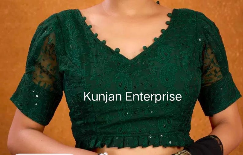 May include: A green embroidered blouse with a V-neckline and short sleeves. The blouse has a ruffled hem and is decorated with small, white buttons. The text "Kunjan Enterprise" is visible on the front of the blouse.