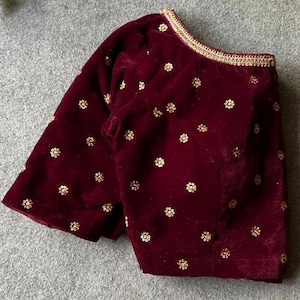 May include: A maroon velvet blouse with gold embroidery. The blouse has a one-shoulder design and a short sleeve.