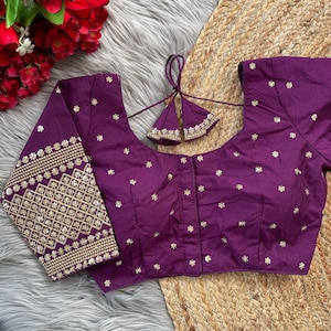 Peut inclure: Un chemisier en soie violet avec des broderies dorées et un nœud décoratif au dos. Le chemisier a un col rond et des manches trois quarts.