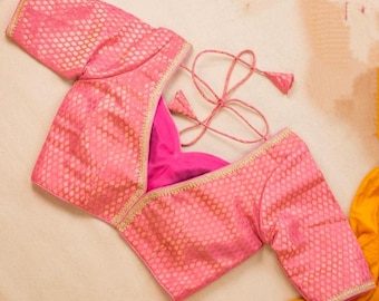 May include: A bright pink blouse featuring a textured diamond pattern and gold-toned trim along the edges. The garment includes short sleeves and a back closure with thin pink fabric ties that end in small, pointed, cone-shaped tassels.