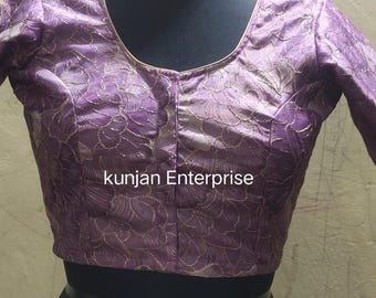 May include: A lavender-colored blouse with long sleeves and a scoop neckline. The blouse features an embroidered floral pattern in a lighter shade of gold. The text "kunjan Enterprise" is printed on the front of the blouse.