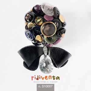 Può includere: Un fiocco in pelle nera con un ciondolo di cristallo pende da un gruppo di bottoni colorati. I bottoni sono di varie forme e dimensioni, tra cui rotondi, quadrati e ovali. I bottoni sono in tonalità di nero, oro, viola e verde. Il ciondolo è un cristallo trasparente con un design sfaccettato. Il testo "rijiventa" e "NUOVE FORME DI PIACERE" è stampato in rosso e nero sotto il fiocco. Il numero "n. S10007" è stampato in nero sotto il testo.