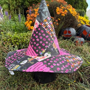 May include: A conical witch hat featuring a patchwork design of black and pink fabric. The pattern includes small pink jack-o'-lanterns, black and white diamond shapes, and various Halloween-themed illustrations like eyeballs and potion bottles.