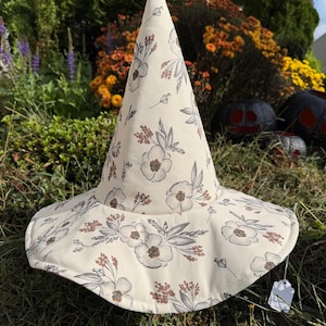 May include: A pointed witch hat made of cream-colored fabric featuring a repeating pattern of gray and rust-colored floral illustrations. The hat has a wide, circular brim and a tall, conical crown, with a small white price tag attached to the edge.