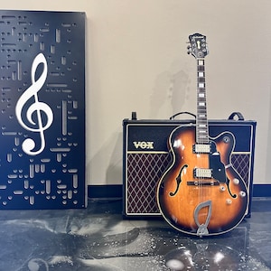May include: A brown and black electric guitar with a black pickguard and a Vox amplifier. The guitar has a tremolo arm and a single cutaway. The amplifier has a black grille with a diamond pattern and the word "Vox" in white letters.
