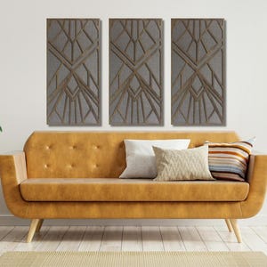 May include: Three abstract geometric wall art panels in a gray and brown color scheme. The panels are arranged in a row on a white wall above a brown leather sofa.