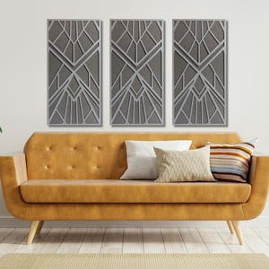 May include: Three gray geometric wall art panels are displayed above a mustard-colored sofa with pillows. The artwork features a layered design of angular shapes. A small wooden side table with plants and a floor rug complete the room's decor.