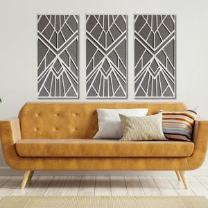 May include: Three gray geometric wall panels with a white geometric design. The panels are hanging on a white wall above a brown leather sofa with pillows.