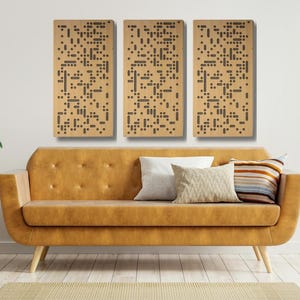May include: Three rectangular wall art panels with a repeating pattern of dark brown dots and dashes. The panels are mounted above a mustard-colored sofa with pillows. The room is decorated with plants and a wooden side table.