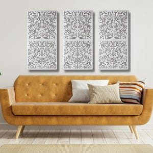 May include: Three white rectangular wall panels with a geometric pattern of small black dots. The panels are hanging on a white wall above a brown leather sofa with two throw pillows. A potted plant is on a wooden stand to the right of the sofa.