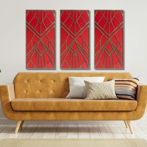 May include: Three red and gold geometric wall art panels hang above a mustard-colored sofa with pillows. The art features a repeating pattern of angled lines. A small side table with a plant and a vase of greenery are on the left.
