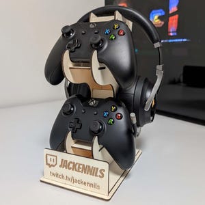 Könnte beinhalten: Ein Holzständer mit zwei schwarzen Videospielcontrollern und einem schwarzen Headset. Der Ständer trägt den Text "JACKENNILS" und "twitch.tv/jackennils".