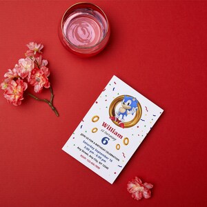 Op de afbeelding: Een verjaardagsuitnodiging met het personage Sonic the Hedgehog. De witte uitnodiging heeft gouden ringen en confetti. De tekst luidt "William wordt 6" met feestdetails. Een glas water en bloemen liggen op een rode ondergrond. Afmetingen zijn niet van toepassing.