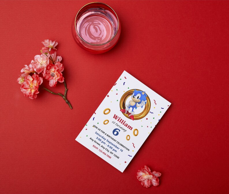 Op de afbeelding: Een verjaardagsuitnodiging met het personage Sonic the Hedgehog. De witte uitnodiging heeft gouden ringen en confetti. De tekst luidt "William wordt 6" met feestdetails. Een glas water en bloemen liggen op een rode ondergrond. Afmetingen zijn niet van toepassing.