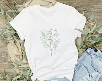May include: White t-shirt with a green line drawing of lily of the valley flowers on the front. The shirt is laying on a wooden surface with a pair of white sneakers and blue jeans.