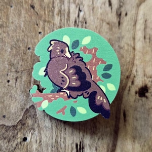 May include: A circular wooden pin with a green background and a brown bird design. The bird is perched on a branch with green leaves. The pin is set against a wooden surface.