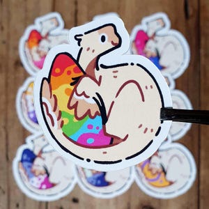 Peut inclure: Plusieurs autocollants représentant un animal de dessin animé embrassant un objet aux couleurs de l'arc-en-ciel. L'animal est beige clair avec des contours noirs et de petits détails. L'objet arc-en-ciel présente des couleurs variées, dont le rouge, l'orange, le jaune, le vert, le bleu et le violet.