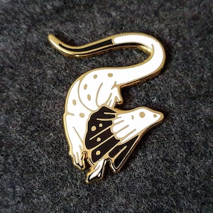 Könnte beinhalten: Ein goldfarbener Metall-Ansteckpin in Form eines stilisierten Tieres mit weißem Körper und schwarzen und goldenen Akzenten. Der Pin hat ein geschwungenes Design und ist wahrscheinlich ein Accessoire.