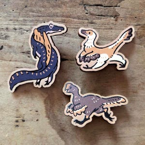 May include: Three wooden dinosaur cutouts. One is a blue and orange Spinosaurus, another is a tan and white raptor, and the third is a purple and tan raptor. The cutouts are on a wooden surface.