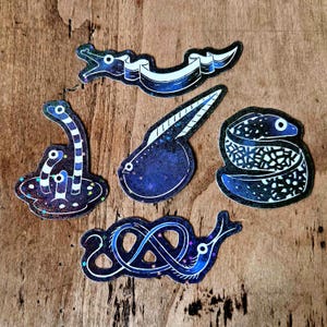 Peut inclure: Un ensemble de cinq autocollants représentant diverses créatures marines. Chaque autocollant a un fond bleu foncé avec des accents blancs et bleus. Les motifs incluent un serpent, un têtard et une créature marine enroulée.