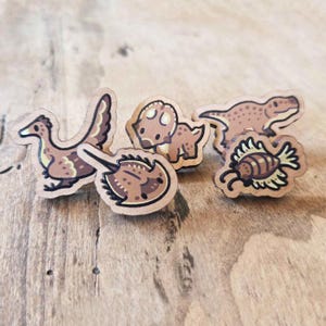 Könnte beinhalten: Ein Set aus vier kleinen Holz-Anstecknadeln mit illustrierten braunen und beigen Kreaturen, darunter eine vogelartige Form, ein Pfeilschwanzkrebs, ein Dinosaurier im Triceratops-Stil und ein kleines insektenartiges Wesen mit Flügeln und dunklen Umrissen.
