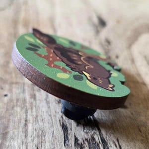 May include: A wooden pin featuring a cartoon illustration of a brown animal on a green background. The pin has a dark brown edge and a black pin backing. The animal is lying down with its head turned.