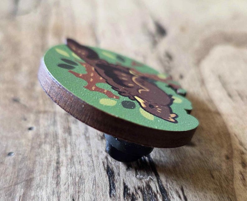 May include: A wooden pin featuring a cartoon illustration of a brown animal on a green background. The pin has a dark brown edge and a black pin backing. The animal is lying down with its head turned.