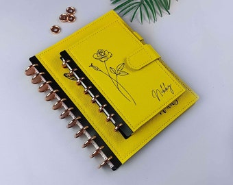 Può includere: Un'agenda in pelle gialla con un design a forma di rosa e il nome "Nikki" inciso sul davanti. L'agenda ha una rilegatura a spirale nera e dorata e una tasca in pelle gialla abbinata.