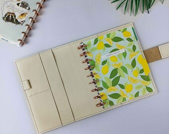 Può includere: Agenda color crema aperta con un inserto a tema limoni e la scritta "Squeeze the Day". L'agenda ha una copertina in pelle, una chiusura a scatto e anelli color oro rosa. È visibile una seconda agenda con un disegno di farfalle.