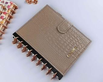May include: A beige faux leather planner with a crocodile embossed pattern. The planner has a black spine with a gold metal disc binding system. The planner has a flap closure with a gold metal button and a gold metal nameplate with the word "Remarkable" engraved on it.