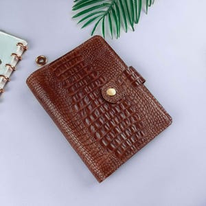 May include: A brown leather journal with a crocodile skin texture. The journal has a flap closure with a gold-coloured button. The journal is closed and appears to be new.