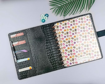Può includere: Agenda in similpelle nera con texture coccodrillo. L'agenda aperta rivela un taccuino a spirale con un motivo a borse della spesa. Sul taccuino c'è la scritta "live creatively". Sono visibili diversi scomparti per carte e una penna.