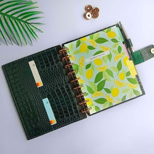 Może przedstawiać: Otwarty ciemnozielony planer z fakturą krokodyla, z wnętrzem w motywie cytrynowym. Planer zawiera długopis, przegródki i uchwyt na karty z dwiema kartami. Tekst "Squeeze the Day" jest widoczny na etykiecie.