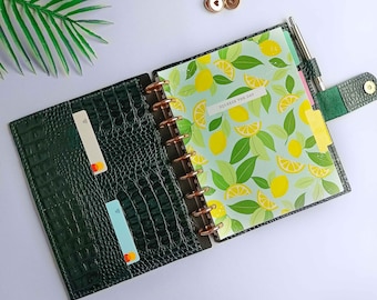 Può includere: Agenda verde scuro con effetto coccodrillo, aperto con un design a tema limoni. L'agenda include una penna, divisori e un porta carte con due carte. Il testo "Squeeze the Day" è visibile su un'etichetta.