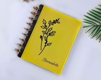 Può includere: Un taccuino rilegato in pelle gialla con un disegno floreale nero e il nome "Bernadette" stampato sulla copertina.