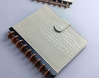 May include: A white faux leather notebook with a crocodile pattern and a rose gold disc binding system. The notebook has a flap closure with a button.