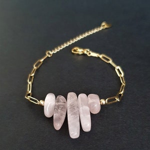 May include: A gold-toned chain bracelet featuring five polished, pale pink, irregular-shaped stone segments arranged in the centre. The bracelet includes a gold-toned metal link chain, a small gold bead on each side of the stones, and an adjustable extender chain with a lobster claw clasp.