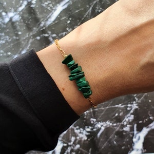 May include: A gold-toned chain bracelet featuring a central cluster of small, irregular, deep green malachite stone chips. The bracelet is secured with a delicate metal chain, showcasing a contrast between the vibrant green mineral and the metallic gold finish of the links.