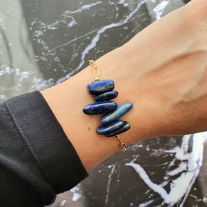 May include: A bracelet featuring five polished, deep blue lapis lazuli stones arranged vertically. The stones are connected by a delicate gold-toned metal chain, creating a linear design that rests against the wrist with a refined and simple aesthetic.