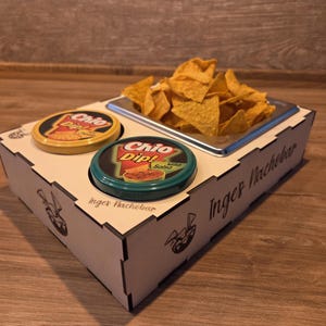 Könnte beinhalten: Ein Holzserviertablett mit einem Metalleinsatz mit Tortilla-Chips. Zwei Behälter mit Chio Dip Salsa befinden sich auf dem Tablett. Auf dem Tablett steht der Text "Inger Nachebar".