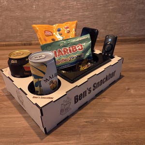Könnte beinhalten: Ein Snack- und Getränke-Organizer aus Holz mit dem Text "Ben's Snackbar". Er enthält zwei Dosen, eine Tüte Chips, eine Tüte Gummibärchen, ein Telefon und eine Fernbedienung. Der Organizer steht auf einer Holzoberfläche.