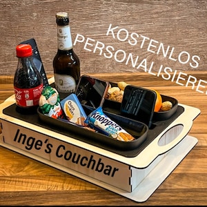 Könnte beinhalten: Ein Couchbar aus Holz mit dem Text "Inge's Couchbar" auf der Vorderseite. Die Couchbar hat ein schwarzes Tablett mit Fächern für Snacks, Getränke und Fernbedienungen. Der Text "KOSTENLOS PERSONALISIEREN" ist im Hintergrund sichtbar.