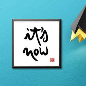 Könnte beinhalten: Ein schwarzes gerahmtes Bild mit den Worten "it's now" in schwarzer Kalligraphie auf weißem Hintergrund. Das Bild hängt an einer blaugrünen Wand über einer gelben Kommode mit zwei Schubladen und einem schwarzen geometrischen Objekt darauf.