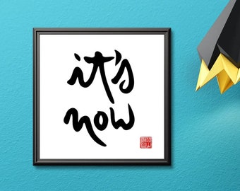 May include: A black framed picture with the words "it's now" in black calligraphy on a white background. The picture is hanging on a teal blue wall above a yellow two-drawer dresser with a black geometric object on top.