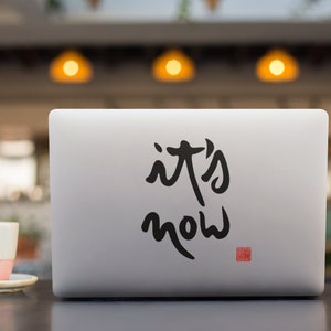 May include: A silver laptop with a black sticker that says "it's now" on the lid. The laptop is sitting on a dark brown table with a white coffee cup and saucer in front of it.