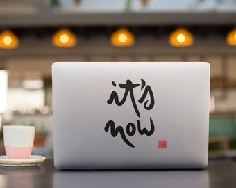 May include: A silver laptop with a black sticker that says "it's now" on the lid. The laptop is sitting on a dark brown table with a white coffee cup and saucer in front of it.