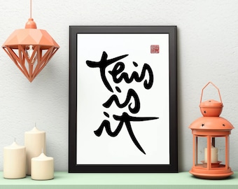 May include: Black and white calligraphy art print in a black frame with the words "This is it" on a white background. The print is hanging on a white wall with a peach-colored diamond-shaped pendant light above it.