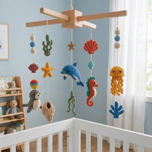 May include: A nursery mobile featuring a wooden cross frame with hanging crocheted sea creatures including a dolphin, seahorse, jellyfish, seal, starfish, and various shells in blue, orange, yellow, and green yarn, suspended by white strings.