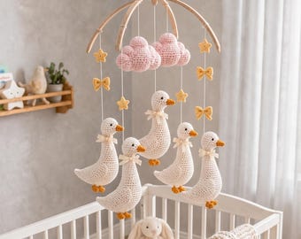 Puede incluir: Un móvil colgante para guardería con cinco patos blancos de ganchillo con picos y patas naranjas, dos nubes rosas de ganchillo, estrellas amarillas y pequeños lazos amarillos suspendidos de un marco de madera curvado con cuerdas blancas.